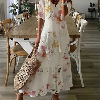 A plus-size model wearing a beige floral chiffon maxi dress with a V-neck, standing next to a houseplant and a chair, showcasing lightweight summer fashion.