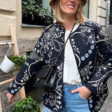 Floral embroidered bohemian paisley jacket worn by a woman smiling, showcasing the detailed embroidery on the sleeves and collar, with a touch of fashion flair. She stands near a window, adding a natural light element to the photograph.