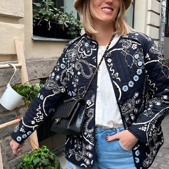 Floral embroidered bohemian paisley jacket worn by a woman smiling, showcasing the detailed embroidery on the sleeves and collar, with a touch of fashion flair. She stands near a window, adding a natural light element to the photograph.