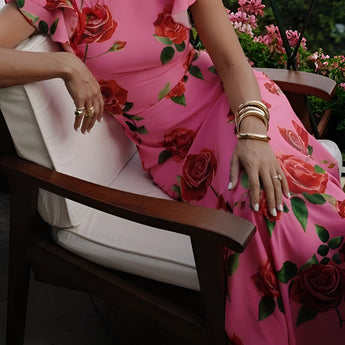 A woman wearing a pink floral mermaid dress with ruffles, suitable for garden parties, smiling as she poses outdoors. She accessorizes with a stylish fedora hat, surrounded by lush greenery, conveying a sense of joy and elegance.