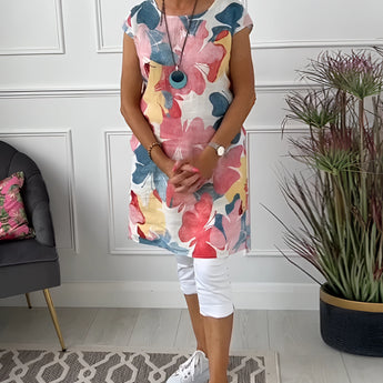 
A woman wearing a floral print sleeveless summer tunic top, standing in a relaxed pose. The top features a vibrant floral pattern with a white background, highlighting the shoulders and arms.