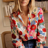 A woman wearing a vibrant floral silk blouse with a spring-inspired design, featuring a variety of colorful flowers. The blouse has a soft, flowing appearance, complementing her casual yet chic fashion style. She is standing in a room with a bookcase in the background, adding a touch of elegance to the setting.