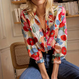 Floral silk blouse in a vibrant spring design hanging on a wooden shelf, featuring a colorful pattern with orange and tartan accents, showcasing its dress shirt style with a focus on the shoulder and neck details.