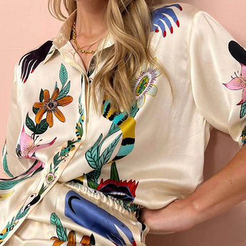 Woman wearing a floral silk blouse with a designer pattern, featuring a collar and long sleeves. The blouse is paired with jeans, and the woman accessorizes with sunglasses.
