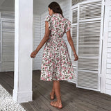A woman wearing a floral summer midi dress with short sleeves and an A-line cut, featuring a tie waist. She is standing in front of a door, showcasing the dress's style.