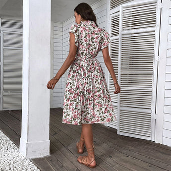 A woman wearing a floral summer midi dress with short sleeves and an A-line cut, featuring a tie waist. She is standing in front of a door, showcasing the dress's style.