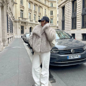 Furry gray oversized women's winter jacket worn by a model in a street style setting, with urban elements like cars and buildings in the background.