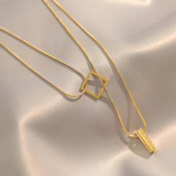 Elegant minimalist necklace with geometric gold bar and square pendant on a delicate chain displayed against a neutral background.