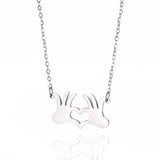 A silver necklace featuring a pendant shaped like hands forming a heart, symbolizing a romantic gesture, placed on a flat surface with decorative plants in the background.