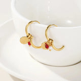 Gold hoop earrings with designer dangle pearl and textured disc charms displayed against a plant backdrop.