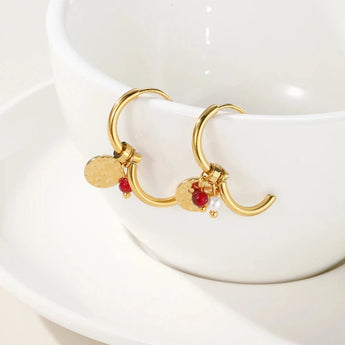 Gold hoop earrings with designer dangle pearl and textured disc charms displayed against a plant backdrop.