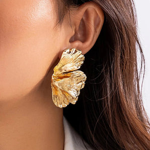 Close-up of a woman's hand holding gold leaf statement earrings with a large textured botanical design.