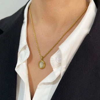 A hand holding a gold locket necklace featuring black onyx and white stone, with the chain draped over a sleeve and blazer.