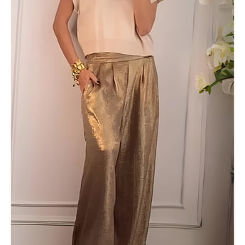 
A person wearing gold pleated metallic trousers, styled for luxury chic party wear, with the focus on the shiny texture of the pants. The trousers are designed with a flattering fit around the waist and fall elegantly towards the ankles. The outfit is complemented by a neutral-toned top and outerwear, set against a minimal background that highlights the clothing's sophisticated appeal.