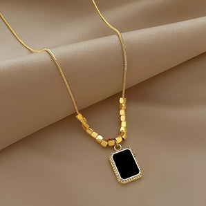 A close-up of a gold snake chain necklace featuring a black rectangular pendant with an adjustable beveled crystal, shown being held by a hand.