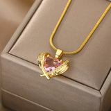 A hand holding a diamond-encrusted heart-shaped pink gemstone gold necklace against a beige wooden background.