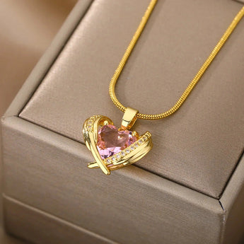 A hand holding a diamond-encrusted heart-shaped pink gemstone gold necklace against a beige wooden background.