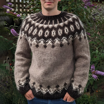 Brown Icelandic wool sweater with a Nordic pattern, featuring long sleeves and a cozy woolen texture, displayed on a mannequin surrounded by grass and plants.
