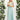 Ivory backless bridesmaid maxi dress with pockets features crossover straps. The dress is styled in light teal with an elegant design, suitable for formal occasions. The model showcases the dress with a focus on its shoulder and neckline details.