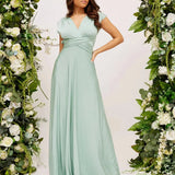 Ivory backless crossover strap bridesmaid maxi dress with pockets, featuring a flowing white fabric and elegant design, displayed on a mannequin surrounded by green plants and white flowers.