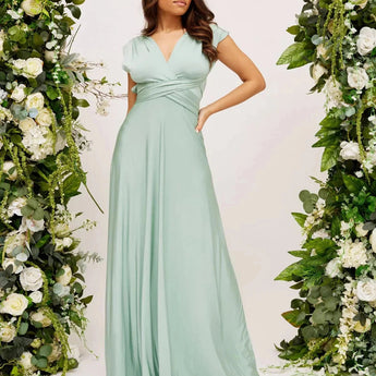 Ivory backless crossover strap bridesmaid maxi dress with pockets, featuring a flowing white fabric and elegant design, displayed on a mannequin surrounded by green plants and white flowers.