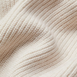 Ivory women's chunky knit cardigan draped over a wooden chair on a hardwood floor with a beige and peach color palette.