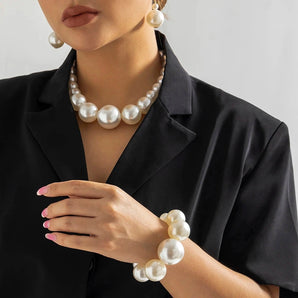 Woman wearing a large pearl jewelry set including a necklace, earrings, and bracelet, with a close-up view of her hand adjusting the necklace; she is dressed in a formal outfit.