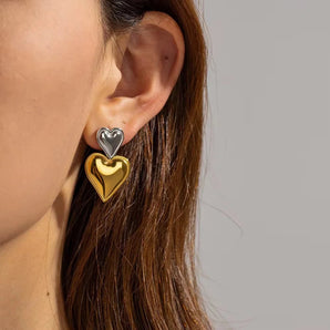 Layered dual-tone heart dangle earrings in silver and gold, shown worn on an ear with visible hair and partial face features like eyebrow and eyelash.