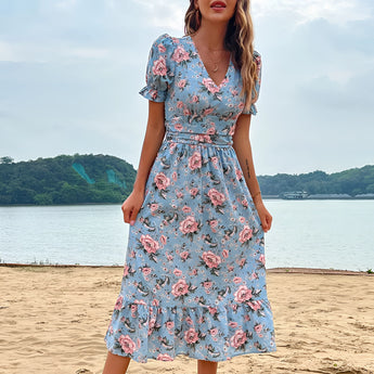 A woman wearing a light blue floral midi dress with a V-neck and short sleeves, standing outdoors near water under a partly cloudy sky.