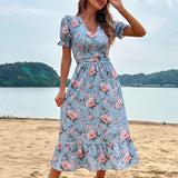 Woman wearing a light blue floral midi dress with a V-neck and short sleeves, standing near a body of water under a clear sky.