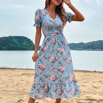 Woman wearing a light blue floral midi dress with a V-neck and short sleeves, standing near a body of water under a clear sky.