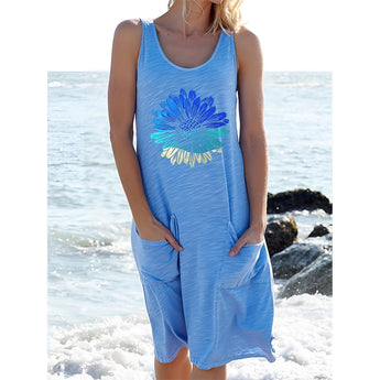 A woman wearing a light blue sleeveless summer dress with a sunflower print, standing on a beach with the ocean in the background.