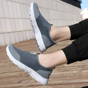 Grey athletic slip-on sneakers designed for walking, featuring a lightweight and breathable material, shown worn on a person's legs.