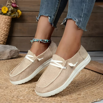A pair of beige linen women's sneakers displayed outdoors, showcasing their eco-friendly and breathable design. The image features the sneakers on a human leg, emphasizing their casual fashion appeal, with a yellow dress and a flowerpot with greenery in the background.