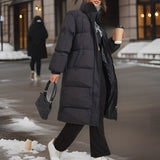 Women's long black insulated winter down coat, featuring a stylish urban design. The coat extends to the knees and includes full-length sleeves, designed for warmth in snowy conditions. It has a waist-cinching fit, resembling a trench coat style, suitable for fashionable outerwear.