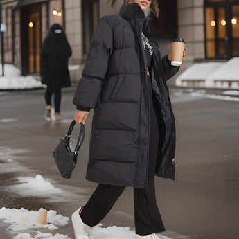 Women's long black insulated winter down coat, featuring a stylish urban design. The coat extends to the knees and includes full-length sleeves, designed for warmth in snowy conditions. It has a waist-cinching fit, resembling a trench coat style, suitable for fashionable outerwear.
