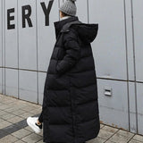 Woman wearing a long black puffer coat with oversized, full-length quilted design, standing on a street, wearing shoes, and a hat with fur trim.