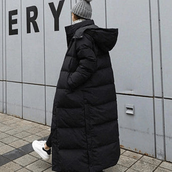 Woman wearing a long black puffer coat with oversized, full-length quilted design, standing on a street, wearing shoes, and a hat with fur trim.