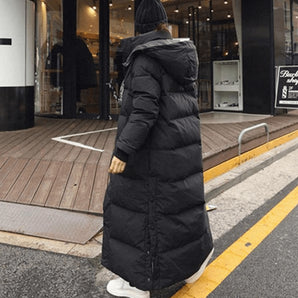Woman in an oversized, full-length black puffer coat with quilted design, standing on an asphalt road.