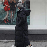 Woman wearing a long black puffer coat, oversized and full-length, standing with a grey hat and carrying a bag.