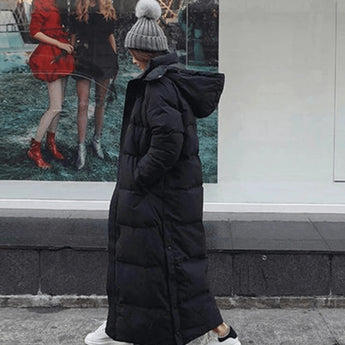 Woman wearing a long black puffer coat, oversized and full-length, standing with a grey hat and carrying a bag.