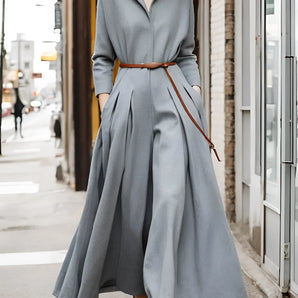 Long grey pleated maxi dress with a belt and collar, showcasing an urban chic design. The dress features long sleeves and a fitted waist, worn by a model with stylish eyewear.