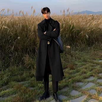 A man wearing a stylish long white double-breasted winter coat, standing outdoors with a clear sky and greenery in the background.