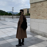 A man wearing a long white double-breasted winter coat and a hat, standing on a street with a cloudy sky and street lights in the background.