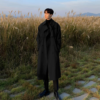 A man wearing a long white double-breasted winter coat with a stylish urban fashion design, standing outdoors against a backdrop of sky and grass.