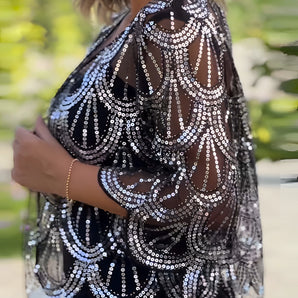 A chic luxury beaded poncho in black with silver accents, draped elegantly over the shoulders. The poncho showcases intricate textile patterns and exudes a sophisticated evening style suitable for formal occasions. The garment is complemented by a tartan-inspired design, enhancing its fashionable appeal.