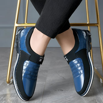 Luxury black leather men's formal shoes with a designer checkered pattern, worn by a person dressed in casual denim clothing, highlighting the sleek and elegant footwear against the lower body.