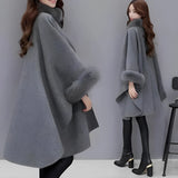 Luxury camel wool cape for women, featuring a faux fur trim around the collar, with a flowing design that drapes over the shoulders and arms, reaching down to the knees. The cape is in a neutral grey tone, with elegant sleeve detailing, ideal for winter wear.