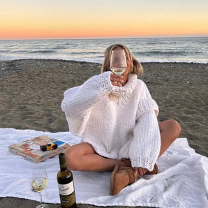 Luxury white wine bottle on a sandy beach during a picturesque sunset, with clear blue skies and gentle waves in the background, evoking a feeling of relaxation and comfort.