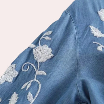 Luxury women's floral embroidered denim bell sleeve top featuring intricate floral designs on the sleeves and body, with a button-up front and a stylish collar, in a shade of grey with hints of electric blue.